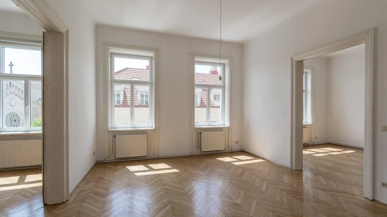 Helles Zimmer mit Fischgrätparkett und hohen Fenstern, das viel Tageslicht hereinlässt.