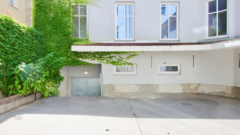 Außenansicht eines Gebäudes mit begrüntem Hof und Zugang zu einem Keller oder einer Tiefgarage.