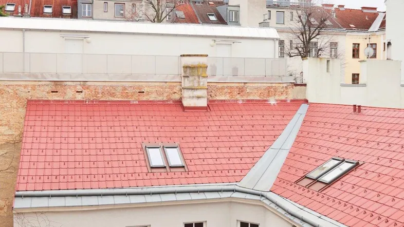 Blick auf die roten Ziegeldächer mehrerer Gebäude mit Dachfenstern und einem Schornstein in städtischer Umgebung.