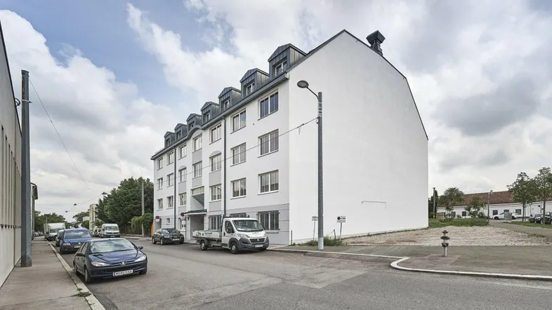 Mehrstöckiges Wohngebäude mit weißer Fassade und Mansarddach an einer Straße mit parkenden Autos.