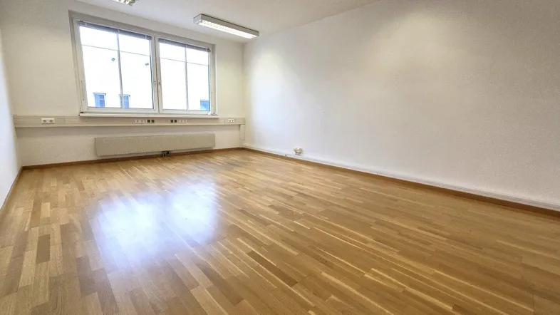 Heller, leerer Büroraum mit glänzendem Holzboden, weißen Wänden und großem Fenster für viel Tageslicht.