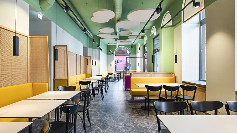 Geräumiger Restaurantbereich mit modernen Tischen, gelben Sitzbänken und grüner Decke, die eine einladende Atmosphäre schafft.