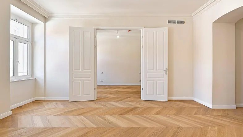 Helles Zimmer mit elegantem Fischgrätparkett, hohen Decken und großen Fenstern, bereit zur individuellen Gestaltung.