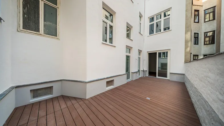 Geräumige Terrasse mit Holzboden und Zugang zu den Innenräumen, umgeben von Gebäudefassaden.