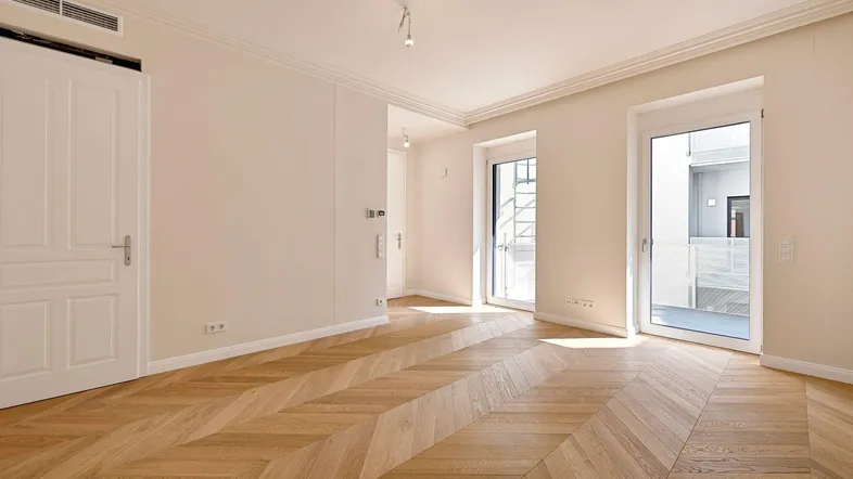Helles Zimmer mit Fischgrätparkettboden und Zugang zu einem Balkon, ideal für Wohn- oder Schlafbereich.