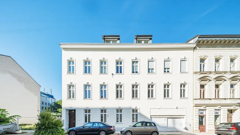 Weißes Mehrfamilienhaus mit klassischer Fassade und Parkplätzen davor unter blauem Himmel.