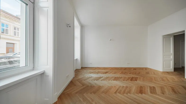 Helles, leeres Wohnzimmer mit Fischgrätparkett und hohen Fenstern, die viel Tageslicht hereinlassen.