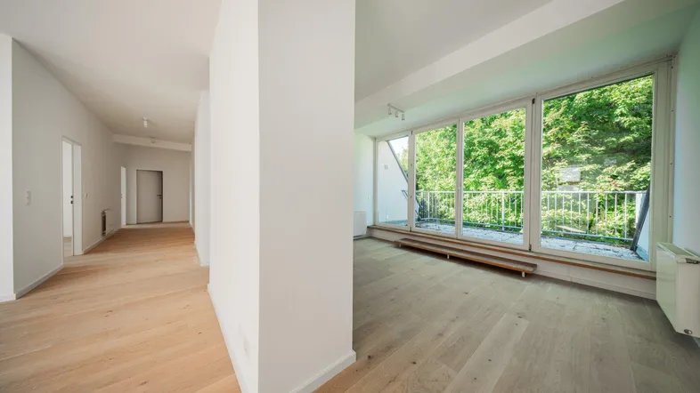 Heller Wohnbereich mit Holzboden und Zugang zu einem Balkon, umgeben von grünen Bäumen.