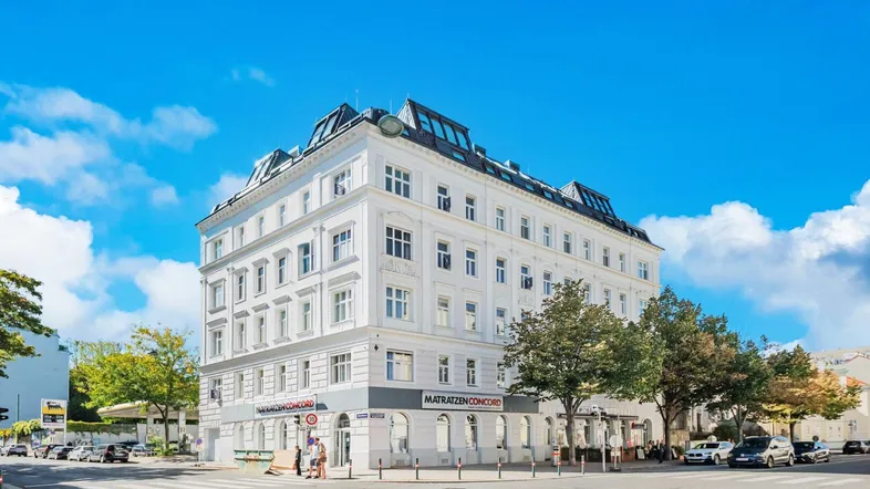 Weißes Eckgebäude mit klassischer Architektur und Geschäften im Erdgeschoss unter blauem Himmel.