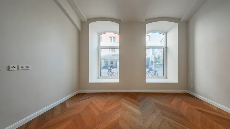 Helles Zimmer mit Fischgrätparkett und zwei Fenstern, die auf die Straße blicken.