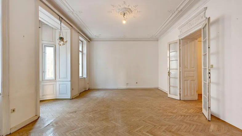 Geräumiges Wohnzimmer mit Fischgrätparkett, hohen Decken und dekorativen Stuckelementen.