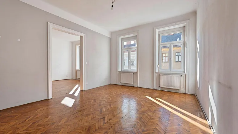 Helles Zimmer mit Fischgrätparkettboden und zwei großen Fenstern, das viel Tageslicht hereinlässt.
