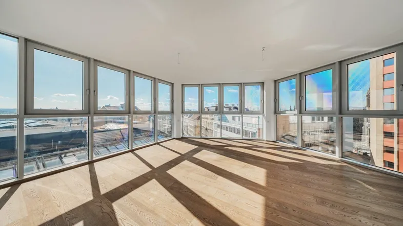 Helles, leeres Zimmer mit vielen Fenstern und Holzboden, das viel Tageslicht hereinlässt.