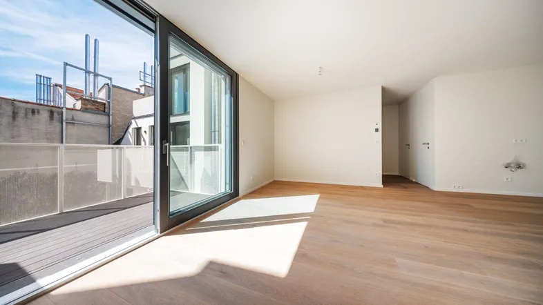 Helles, leeres Zimmer mit Holzboden und großen Schiebetüren, die zu einem Balkon führen.
