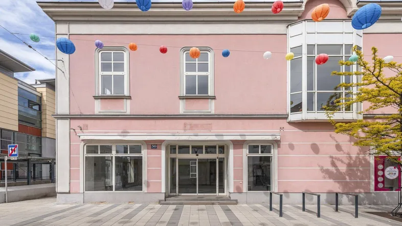 Rosa Gebäude mit großen Schaufenstern und einem Erker, geschmückt mit bunten Laternen an der Straße.
