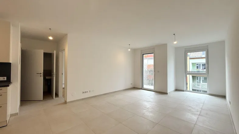 Heller, leerer Wohnbereich mit großen Fliesen, weißen Wänden und Zugang zu einem französischen Balkon.