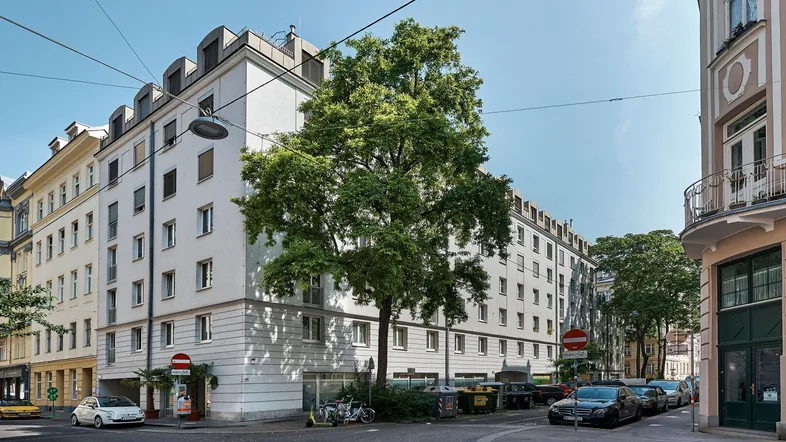 Mehrstöckiges Wohngebäude an einer Straßenecke mit Bäumen und parkenden Autos unter blauem Himmel.