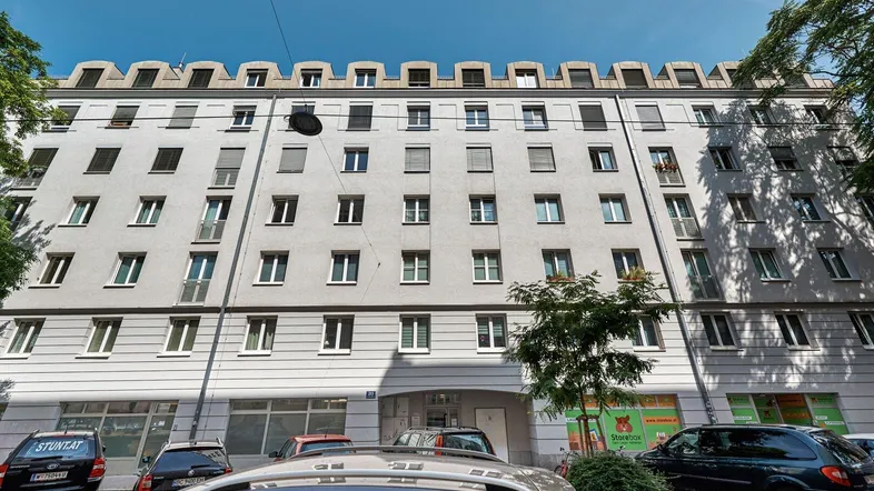 Gebäudefassade mit zahlreichen Fenstern und Rollläden, davor parkende Autos auf der Straße.