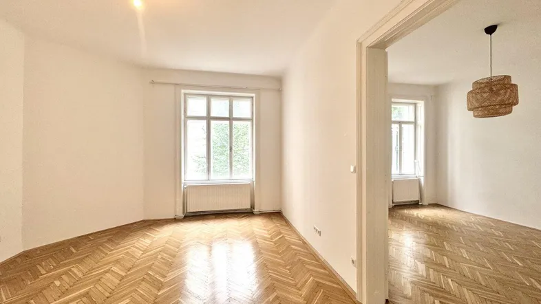 Heller Wohnraum mit glänzendem Fischgrätparkett und hohen Decken, der in einen weiteren Raum führt.