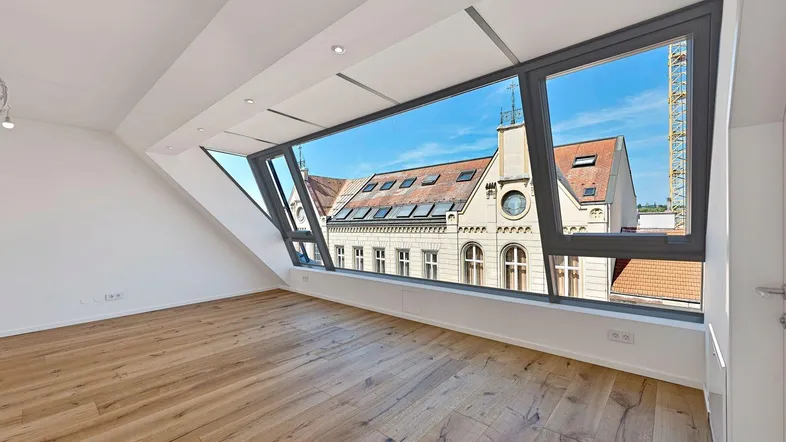 Helles Dachgeschosszimmer mit großen Fenstern, Holzboden und weitem Blick auf die Stadt.