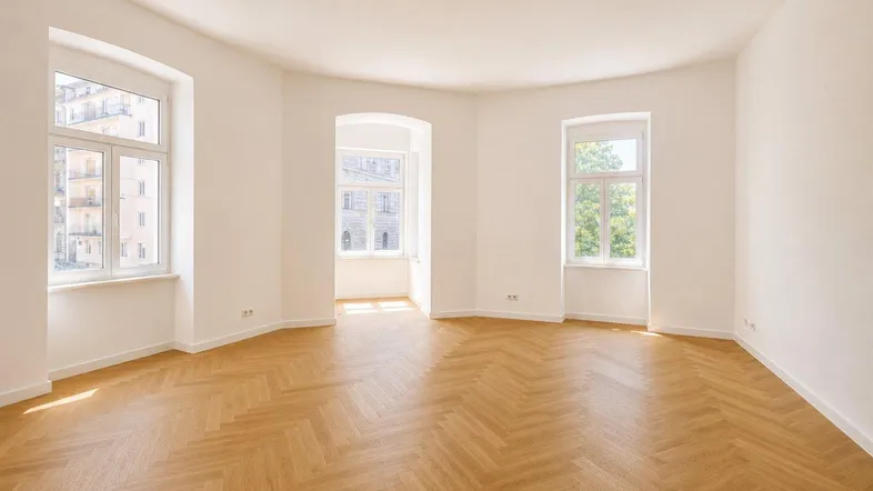 Helles, leeres Zimmer mit Fischgrätparkett und mehreren Fenstern, die viel Tageslicht hereinlassen.