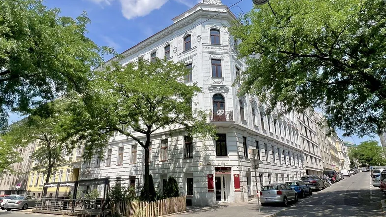 Historisches Eckgebäude mit weißer Fassade und Bäumen an einer städtischen Straße.