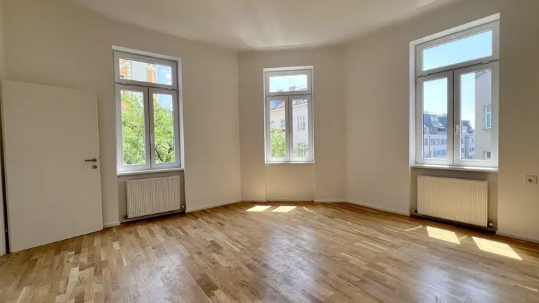 Helles Zimmer mit Parkettboden und mehreren Fenstern, das viel Tageslicht hereinlässt.