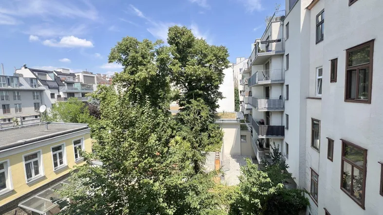 Blick von einem Balkon oder Fenster auf Bäume und umliegende Gebäude unter blauem Himmel.