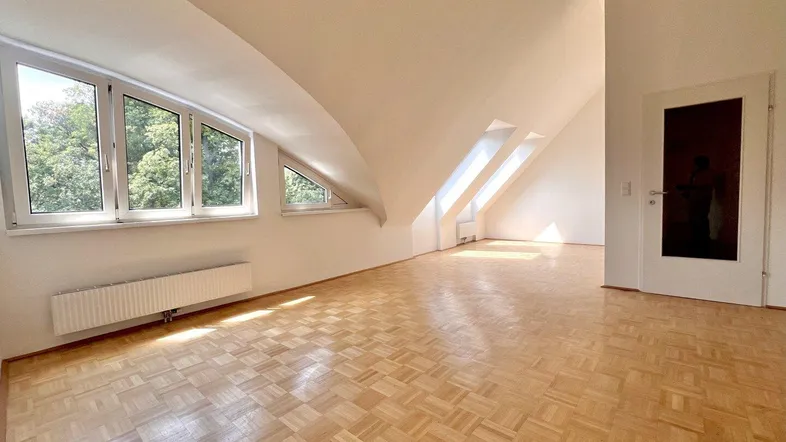 Helles Dachgeschosszimmer mit Parkettboden, schrägen Wänden und mehreren Fenstern, die viel Tageslicht hereinlassen.