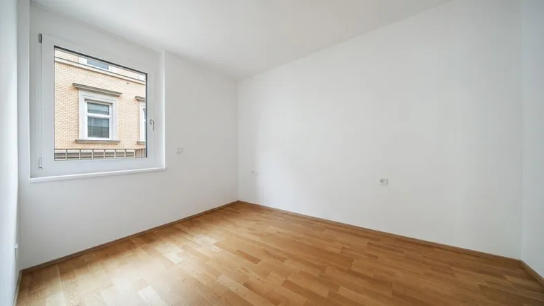 Heller Raum mit weißen Wänden und Parkettboden, mit Blick auf ein gegenüberliegendes Gebäude.