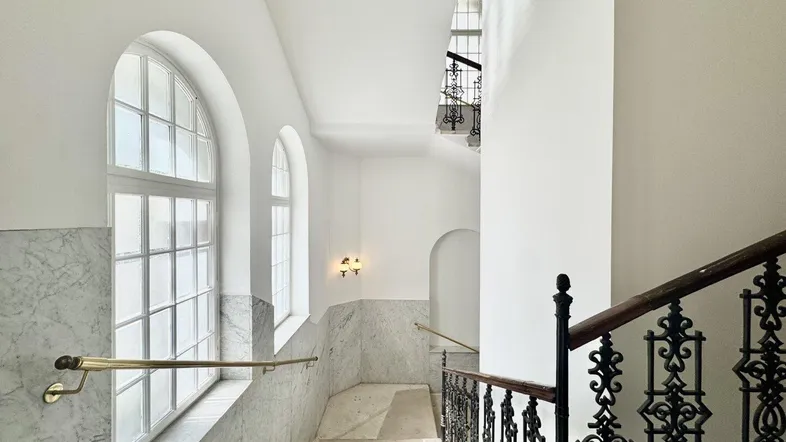 Helles Treppenhaus mit eleganten Marmorwänden, schmiedeeisernem Geländer und großen Bogenfenstern.