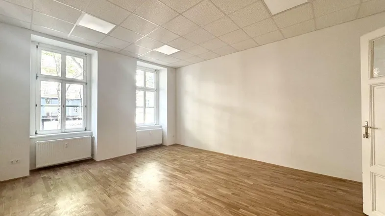 Geräumiger, leerer Raum mit Holzboden und zwei großen Fenstern, ideal für Büronutzung.