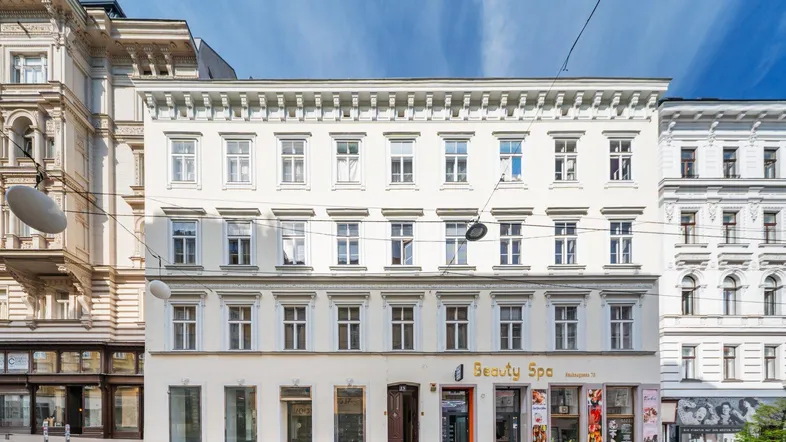 Weißes Mehrfamilienhaus mit mehreren Etagen und Geschäften im Erdgeschoss an einer Stadtstraße.