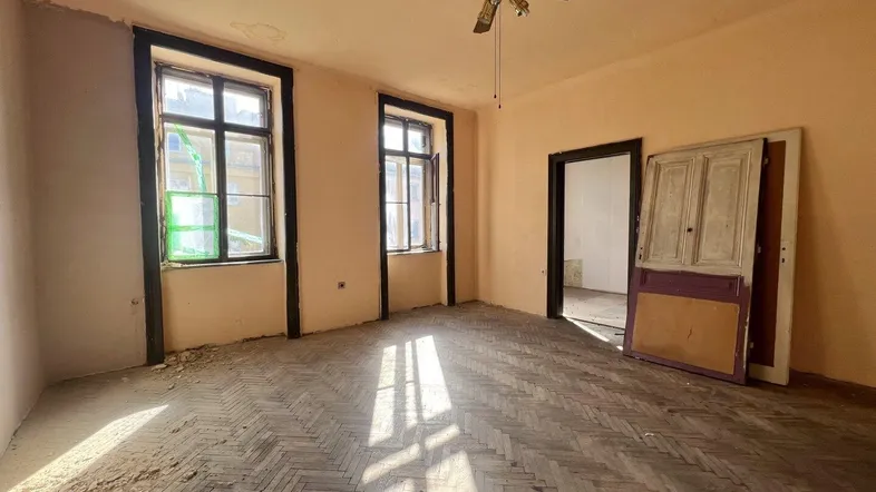 Geräumiges Zimmer mit Fischgrätparkett, hohen Fenstern und Renovierungsbedarf, beleuchtet von Tageslicht.