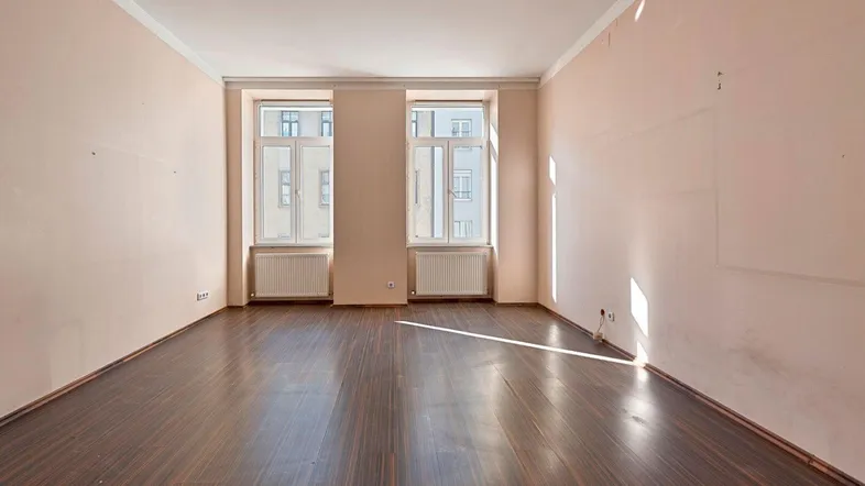 Helles, leeres Zimmer mit zwei Fenstern, Parkettboden und hellbeigen Wänden.