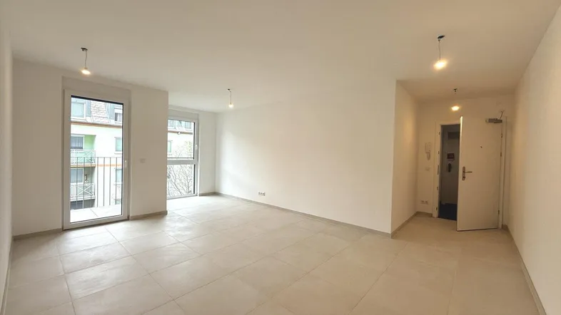 Heller, leerer Wohnbereich mit Fliesenboden, weißen Wänden und Zugang zu einem Balkon.