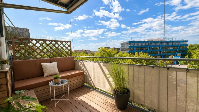 Gemütlicher Balkon mit Holzboden, Sitzbank und Blick auf die Stadt unter blauem Himmel.