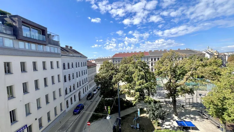 Blick auf eine belebte Straße mit Gebäuden, Bäumen und einem kleinen Parkbereich unter blauem Himmel.