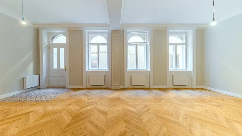Geräumiges Wohnzimmer mit Fischgrätparkett, hohen Decken und großen Fenstern, die viel Tageslicht hereinlassen.