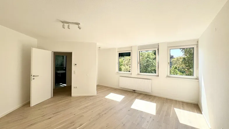 Helles, leeres Zimmer mit Holzfußboden und Blick ins Grüne durch drei Fenster.
