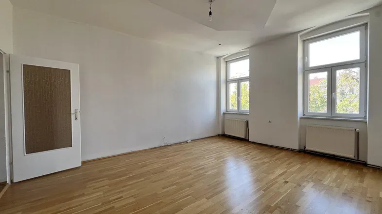 Helles, leeres Zimmer mit Parkettboden und zwei großen Fenstern, bereit zur individuellen Gestaltung.