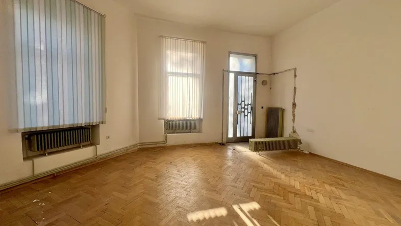 Helles Zimmer mit Fischgrätparkett, Heizkörpern und Fenster mit Jalousien.