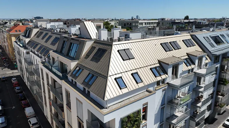 Luftaufnahme eines modernen Mehrfamilienhauses mit weißer Fassade, Mansarddach, zahlreichen Balkonen und Photovoltaikanlage.
