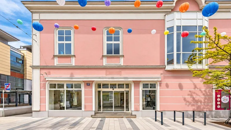 Rosa Gebäude mit großen Fenstern und bunten Laternen, das eine lebendige Straßenansicht bietet.