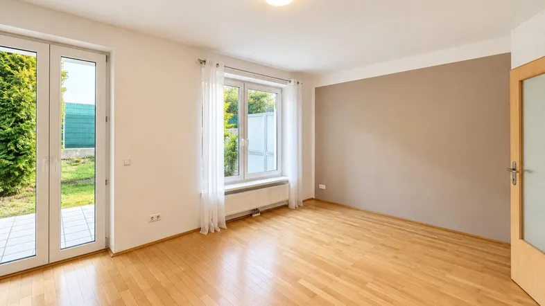 Helles Zimmer mit Holzboden, großem Fenster und Zugang zu einer kleinen Terrasse mit Garten.