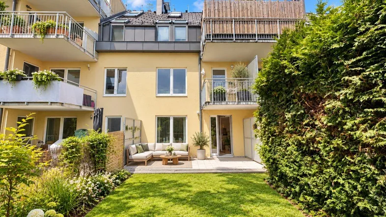 Gepflegter Garten mit Rasenfläche und möblierter Terrasse hinter dem Wohngebäude unter blauem Himmel.