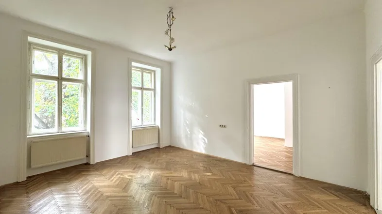 Helles Zimmer mit Fischgrätparkett und zwei großen Fenstern, das viel Tageslicht hereinlässt.