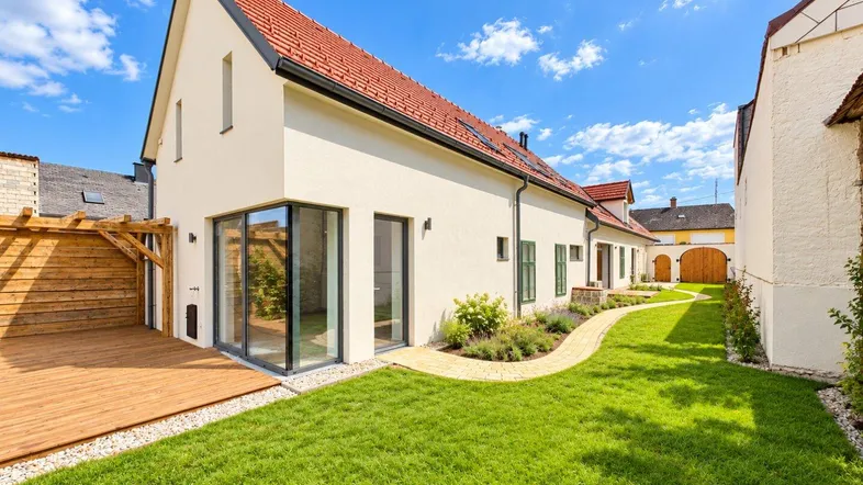 Modernes Haus mit großem Garten, Holzterrasse und gepflegter Rasenfläche unter blauem Himmel.