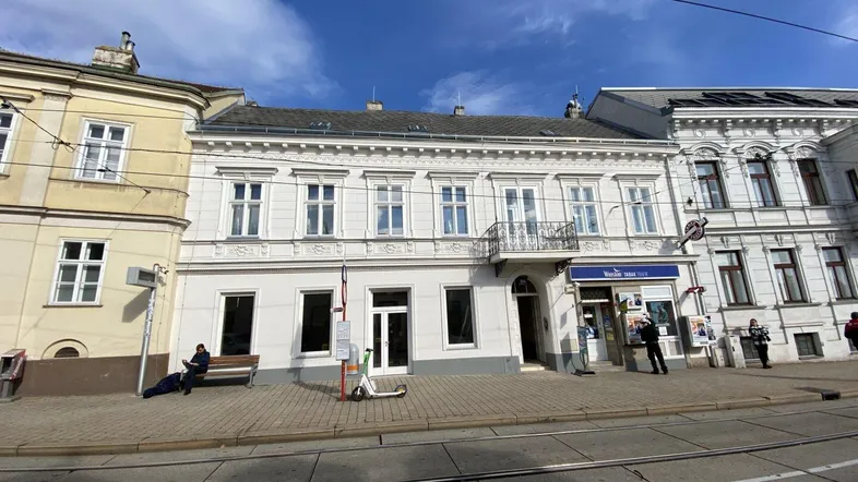 Historisches Gebäude mit klassischer Fassade, kleinem Balkon und Geschäften im Erdgeschoss an einer belebten Straße.