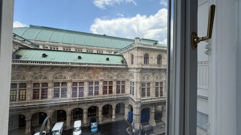 Blick aus einem Fenster auf die Wiener Staatsoper und die Straße mit parkenden Fahrzeugen.